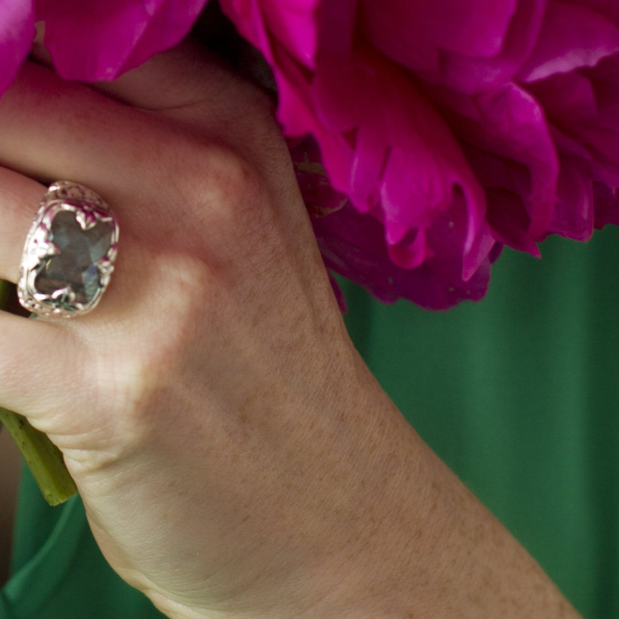 Silver ring featuring a cushion-cut labradorite gemstone with an iridescent blue-green flash, set with elegant leaf-like prongs and an intricately carved filigree scrolling band
