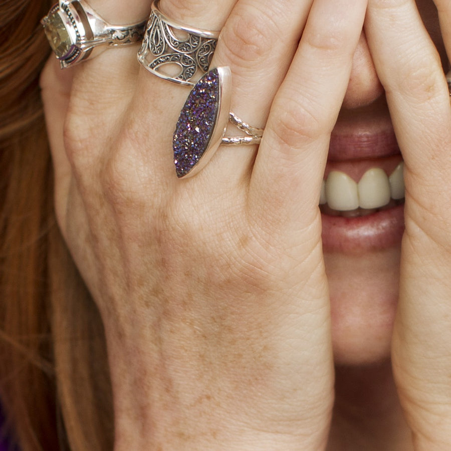 Photograph of a silver ring featuring a large oval iridescent purple druzy gemstone sparkling with blue and gold flecks and a hammered textured band on a white background